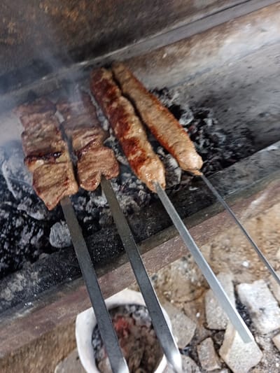 The image shows three skewers of grilled meat cooking over charcoal. The first two skewers on the left feature chunks of meat, appearing well-done. The two skewers on the right contain elongated meat products, similar to sausages or kebabs. They exhibit a reddish-brown color. The grilling setup seems to be an outdoor or makeshift arrangement, with materials like stone and brick visible in the foreground. Smoke is present, indicating active cooking.
