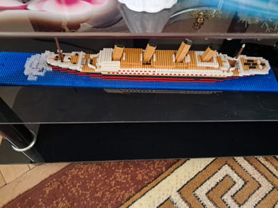 The image displays a detailed model of the Titanic, meticulously constructed from small building blocks, akin to Lego. The ship features a white superstructure, a tan deck, and a distinctive hull with red, black, and white stripes. Four tan and black smokestacks rise from the deck, and a mast is visible at the bow.
The model is placed on a vibrant blue studded base plate, simulating water. At the ship's bow, translucent white blocks are arranged to depict waves, suggesting movement through water or possibly an impact, referencing the ship's fate.
The entire setup rests on a dark, reflective surface, likely a shelf or table. Above this, a blurry reflection of an indoor environment is visible on what appears to be a glass tabletop or shelf above the model, showing soft pink and white elements, possibly from decorative objects or lighting. Below the dark surface, a section of a patterned rug or carpet is visible, featuring a geometric design in shades of brown, tan, and cream. A metallic, curved leg of furniture is also discernible in one of the crop images, supporting the dark surface.
There are no people present, and no specific activity beyond the static display of the model. The setting is indoors, with no discernible time of day or weather conditions. There is no visible text in the image. The scene suggests a decorative display within a home or similar indoor space in Gavar, Armenia.