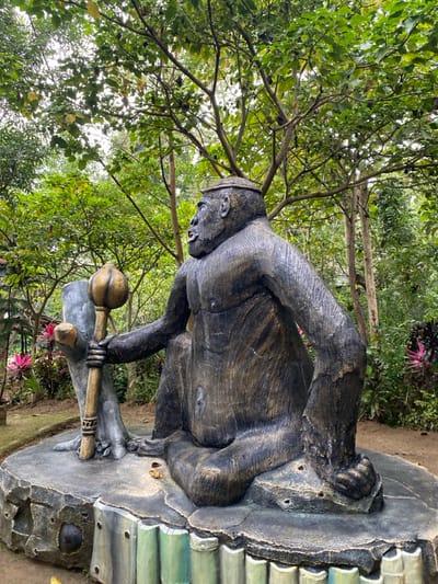 A bronze statue of a gorilla, seated and holding a staff topped with a lotus flower, sits on a rocky platform with a decorative base in the forest. The statue is life-sized and the gorilla is facing the right side of the frame. The statue appears to be located in a lush green forested area in Kecamatan Manyaran, Indonesia (latitude: -7.826, longitude: 110.816).