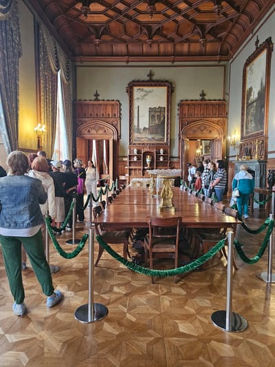Captured inside Vorontsov Palace, located in Алупкинский городской совет, Ukraine, a group of tourists are observing a long table displaying various objects. The table, positioned in the center of the room, is cordoned off by stanchions connected by green velvet ropes. Several tourists, some wearing protective shoe coverings, are visible on both sides of the stanchions. The room features an intricately patterned wooden ceiling and parquet flooring. Ornately carved wooden panels line the walls, adorned with framed paintings. The lighting is a mix of natural and artificial illumination from chandeliers.