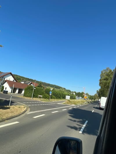 The image shows a clear blue sky over a residential area with a road in the foreground. On the left side of the road, there are houses with red-tiled roofs and solar panels. There are also green trees and bushes. On the right side of the road, there are more trees and a white van. The road has white dashed lines indicating lanes and a pedestrian crossing. In the distance, a construction crane is visible. The scene appears to be a sunny day in a suburban area. Based on visual cues and information from similar scenes, this could be a location in Wartenberg, Germany.