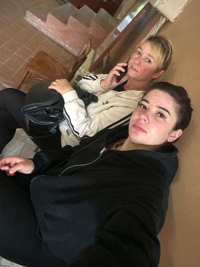 In the city of Gavrilov Posad, Russia, two individuals are seated on a step near a tiled floor and a light brown wall. The person on the left, a woman with short blonde hair, is holding a black smartphone to her ear, engaged in a conversation. She is wearing a white zip-up hoodie with black stripes on the sleeves. Beside her, a younger woman with dark hair is also seated, wearing a black zip-up hoodie. She is looking directly at the camera. In the background, a wooden stool and a section of stairs are visible.
