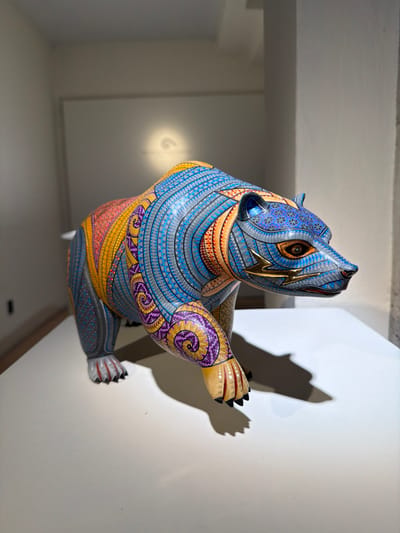 A vibrant, hand-painted wooden bear statue is positioned in a corner. Its body is adorned with intricate patterns in shades of blue, gold, purple, and orange. The head features golden accents, with detailed eyes and ears. The statue's shadow is cast on the white wall, creating a distinct contrast. The surrounding environment includes sections of a bright, white wall and a dimly lit ceiling, emphasizing the bear's colorful presence in the space.