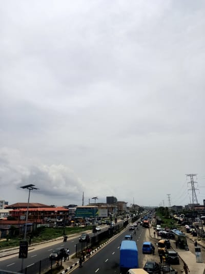 The image captures a wide-angle view of a bustling multi-lane highway or major road in Eputu Town, Nigeria, under an overcast sky. The scene depicts a busy urban landscape with significant vehicle and pedestrian traffic.

In the foreground and midground, a divided road stretches into the distance, flanked by buildings and various activities. The road itself shows cars, buses (including a prominent blue bus on the left lane), and motorcycles in motion, indicating active traffic flow. Pedestrians are visible walking along the shoulders and median barriers, as well as crossing the road.

On the left side of the road, multi-story buildings with reddish-brown roofs line the street, interspersed with patches of green vegetation. Modern-looking streetlights equipped with solar panels are mounted on tall poles. A large billboard is visible with text, including "Zoho.com" and phrases like "Complexity ends NOW. Scale smart with Zoho."

The right side of the image reveals a more informal commercial area adjacent to the road. Numerous people are gathered around makeshift stalls and parked vehicles, suggesting street vending or market activity. Large electricity pylons, carrying high-voltage transmission lines, are prominent in the right background, extending into the distance and indicating significant infrastructure.

The weather appears to be overcast, with a predominantly grey, cloudy sky that diffuses the daylight. The time of day is clearly daytime, but without direct sunlight. The overall impression is one of a vibrant, active urban thoroughfare characteristic of a rapidly developing city in Nigeria.