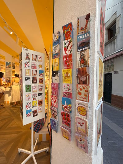 This media file shows an outdoor market stall, likely in Tossa de Mar, Spain, as indicated by the text "TOSSA" and "COSTA BRAVA" on some of the displayed items. The stall is filled with colorful postcards and prints, many featuring food items like paella, tomatoes, and churros, along with Spanish phrases and cultural references. The scene appears to be set during the daytime, with bright lighting suggesting fair weather.
Two people are visible in the foreground, appearing to be a customer and a vendor interacting at the stall. The customer, wearing a light-colored dress, is looking at the items on display, while the vendor is partially obscured but seems to be assisting. The background shows a street with buildings, typical of a European town. The overall atmosphere seems relaxed and casual, characteristic of a tourist-oriented market. The prominent display of various printed materials suggests this is a place selling souvenirs or local art. Several of the items have prices listed, such as "7,50€". The vibrant colors and the array of products create a lively and inviting scene.