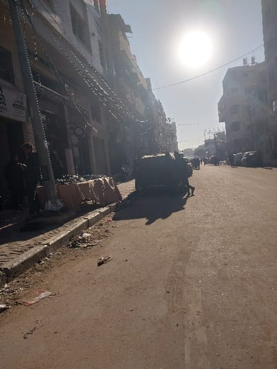 This image depicts a street scene in a-Zawayda, Palestinian Territory, under bright daylight with a prominent sun causing strong backlighting and lens flare. The worn, dusty road stretches into the distance, populated by multiple pedestrians and several indistinct vehicles. Multi-story buildings with somewhat deteriorated facades line both sides of the street; those on the left feature ground-level storefronts. A dense network of overhead electrical or communication cables is visible, strung between utility poles and across the street. On the left sidewalk, a temporary vendor setup consists of a table covered with a light-brown cloth, displaying dark, piled goods. Two individuals are standing near this display, one a man with dark hair wearing a dark jacket. Scattered debris is visible along the curb and on the sidewalk. In the mid-ground, a large, dark, undefined object, possibly a vehicle or cart, casts a prominent shadow across the road.