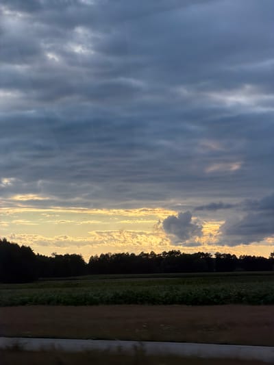 This is a full shot of a cloudy sky over a rural landscape in Kopaniny, Poland. The sky is filled with layers of dark gray and blue clouds, with some lighter patches of yellow and white peeking through, suggesting either sunrise or sunset. Below the sky, a dark line of trees runs horizontally across the image, forming the horizon. In front of the trees, a field of green crops extends forward, and further down, a strip of brown land is visible, possibly a dirt road or another cultivated field. The overall impression is one of a serene, natural setting under a dramatic sky.