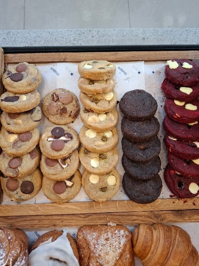 An overhead view captures a commercial baked goods display in Bnei Dror, Israel. The upper portion features a light-colored wooden tray containing multiple vertical stacks of cookies, resting on white paper liners branded with "SPARROWS" and a small diamond pattern. From left to right, the cookie varieties include: a stack of tan cookies with milk and dark chocolate chips; a cluster of tan cookies, some with various chocolate chips and possibly marshmallow-like toppings; a stack of tan cookies with white chocolate chunks and green specks (likely pistachios); a stack of dark brown, unadorned chocolate cookies; and a stack of red velvet cookies, each topped with a dollop of white cream cheese frosting and some containing dark fruit. Below this tray, a second display level shows additional pastries: a dark-colored pastry partially visible on the far left, a white-glazed sweet roll, a square golden-brown pastry topped with streusel, and a classic light brown croissant on the far right. The display rests on a light-colored counter with a dark gray or black edge.