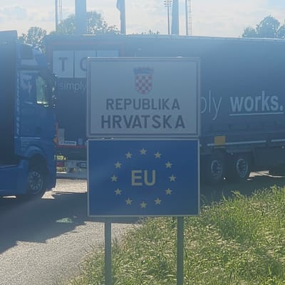 The image shows a border crossing between Croatia and Serbia. The signs indicate that the image was taken at the border between the Republic of Croatia and the European Union. The Republic of Croatia sign includes the Croatian coat of arms. The EU sign contains the EU flag with 12 stars and the letters "EU" in the center. The location is within the general vicinity of Sid Municipality, Serbia. A truck is partially visible in the foreground and background. Grass can be seen to the right of the sign.