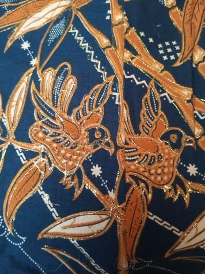 Dark blue fabric displays a repeating pattern of stylized brown birds facing each other. The birds have intricately detailed wings and bodies. Bamboo stalks, also in brown, weave through the design. Small white stars and geometric shapes fill the spaces between the primary motifs. A shimmering gold outline highlights the edges of the birds, bamboo, and other decorative elements, adding a touch of elegance to the fabric.
