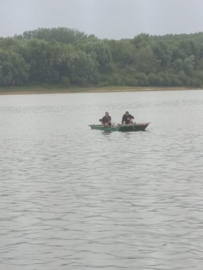 Two people are in a small boat on a body of water. The boat is green and has a red stripe along the top edge. One person is sitting in the front of the boat and the other is in the back. They are both wearing dark clothing. The water is rippled, suggesting it is moving. In the background, there is a shoreline with trees. The trees are dense and green, forming a thick line along the water's edge. The sky is overcast and appears hazy or foggy. The overall scene conveys a sense of a calm, possibly cool day. The location is identified as Šabac, Serbia. No text is visible in the image.