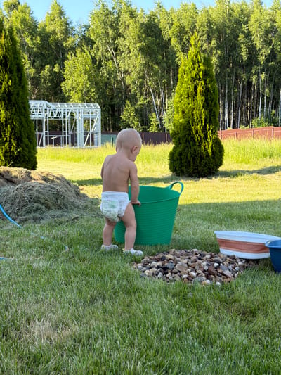 In Борисьево Днп, Russia, a young child wearing a diaper and sandals stands in a grassy yard. The child faces away from the camera, looking towards a large green bucket filled with pebbles. To the child's left, a pile of hay is visible, and a blue hose is coiled on the grass. In the background, a partially constructed greenhouse with a metal frame stands amongst tall trees. A wooden fence runs along the edge of the property. Two evergreen trees are positioned in the foreground, one on the left and one on the right of the child.