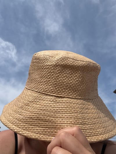 A close-up, eye-level shot shows a person's head and shoulders obscured by a woven straw hat, against a backdrop of a cloudy blue sky. The hat is wide-brimmed and bucket-shaped, with a textured weave. A person's hand, with fingernails painted pink, is visible holding the brim of the hat near their face. The individual is wearing a black spaghetti strap top. The scene appears to be outdoors, possibly in a park or beach setting. There is no specific indication of the city of Ayyrtau, Kazakhstan in the image.