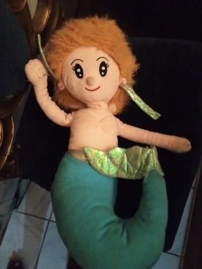 A plush mermaid doll with bright orange hair, rosy cheeks, and large, expressive eyes rests on a dark surface. She has light pink skin and a green mermaid tail. A shimmering, green, leaf-like fabric adorns her waist. Her arms are outstretched, and she has green, streamer-like earrings. The doll appears to be handmade or of simple construction. A partially visible chair with metallic gold trim is situated near the doll.