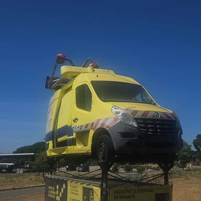 This media file captures a bright, sunny day with a clear blue sky, likely in the afternoon. The main subject is a yellow service van, possibly a Renault Master, prominently displayed on a raised platform. The van appears to be an older model, showing some signs of wear and tear, including a cracked windshield and a missing hubcap. It's equipped with emergency lights on its roof and reflective markings on the front and sides. The van is positioned outdoors, with a grassy area and trees in the background. In the distance, there's a gas station with a canopy and several cars, suggesting a public roadside location.
A key element of the scene is the signage displayed on the fencing below the van. These yellow banners feature text in French, emphasizing safety. One sign reads "RESPECTEZ LE CORRIDOR DE SÉCURITÉ," meaning "Respect the safety corridor." Another sign, partially visible, seems to provide instructions related to approaching an intervention vehicle. This context, combined with the van's markings (potentially indicating it's from a service or maintenance company, "ASF" is visible), suggests this is an educational display or a safety awareness installation. There are no people actively engaged in any specific activity; the scene appears static, focusing on the displayed van and safety information. The overall impression is one of a warm, summery day used to highlight road safety messages.