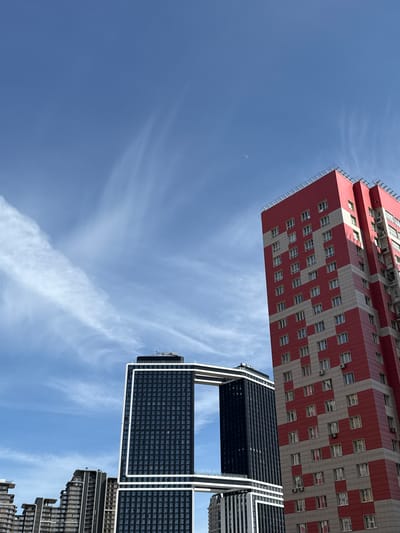 This image captures a unique architectural vista in Perepechino, Russia, under a bright, clear blue sky adorned with wispy white clouds. The photograph is taken from a low, upward-looking perspective, showcasing two distinct modern buildings.
On the left, a tall building features a striking facade with a repeating pattern of red and light-colored (possibly white or light grey) rectangular blocks, resembling a checkerboard. Numerous windows are visible across its surface, along with some railings or balconies on its sides.
To the right, a contrasting building with a dark, almost black, facade and prominent white structural outlines dominates. This building appears to be composed of multiple segments or blocks, with a distinctive grid pattern on its dark panels, suggesting a contemporary design. Below it, partially visible, is another building that shares a similar dark aesthetic, contributing to a modern urban landscape.
The sky, occupying a large portion of the frame, is a vibrant blue, indicating daytime. Faint, streaky clouds stretch across the upper parts of the sky, and a subtle crescent moon is also visible. There are no people or specific activities depicted in the scene, and no discernible text is present on the buildings or in the sky. The image emphasizes the architectural forms and their interplay with the expansive sky.