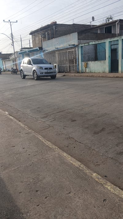 The image shows a street in Barcelona, Venezuela, with a two-story building on the left and a sidewalk on the right. Two cars, one silver and one white, are parked along the building. A motorcycle is parked behind the white car. Power lines run overhead. The building is painted blue and has barred windows and a metal gate. The street is paved and shows signs of wear. The sidewalk is concrete.
