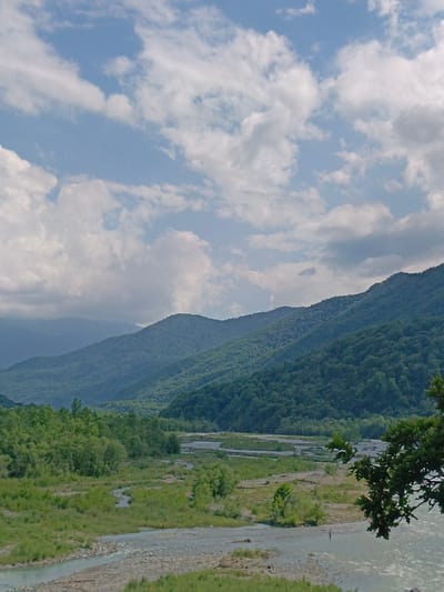 The image shows a wide river flowing through a valley with lush green vegetation on either side. In the background, several layers of mountains rise, covered in dense forests. The sky is blue with scattered white clouds. The riverbed appears stony and the water is clear. The scene is bathed in natural daylight, suggesting it was taken during the day. There is a single person visible in the river, appearing to stand or wade.