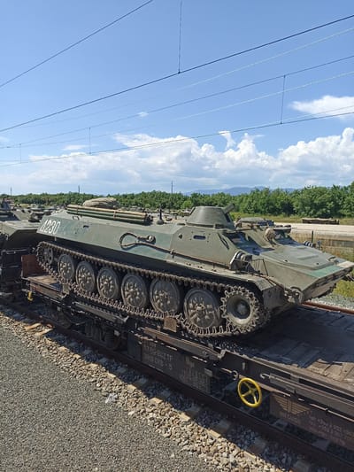 A green, tracked, armored personnel carrier (APC) is secured on a flatbed railcar. The APC has the number "4230" painted on its side. The flatbed railcar has markings and information printed on it, including "3T RIV," "52 BG-BDZ TP," "3919 066-5," "Rgs," "TAIBICI 15140-148-156," "120t - 009," "BDZ CARGO," and contact information for BDZ Cargo. The railcar also features a bright yellow wheel-shaped mechanism on its side. The image is taken outdoors on a sunny day with a blue sky and scattered white clouds. In the background, there is a tree line and possibly some low mountains in the distance. Gravel surrounds the railroad tracks. While the location is suggested to be Zimnitsa, Bulgaria, there are no identifying features in the image itself to confirm this.