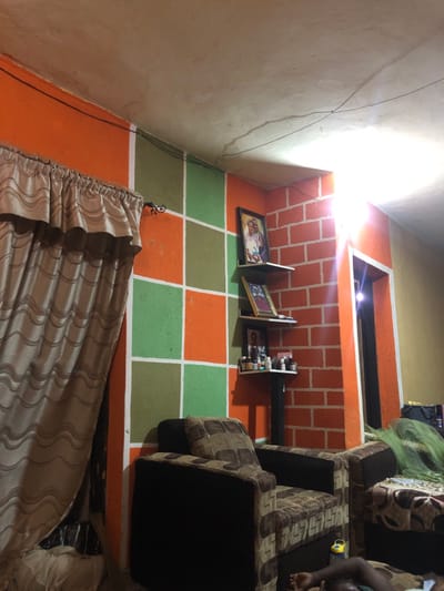 An interior shows a checkered floor in green and orange. A couch with brown patterns sits to the right, partially obscuring the view. The wall behind the couch is orange, styled with a brick pattern and shelves holding pictures and items. A window sits atop the wall. A curtain hangs at the bottom, and bare wires run up the left wall.