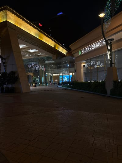 This is an outdoor night scene in Petaling Jaya, Malaysia. The foreground shows a paved walkway with interlocking pavers. A Shake Shack restaurant is visible on the left. Beyond Shake Shack, the facade of a building with intricate yellow illuminated detail is present. Further into the scene, the Coach store is noticeable. People are visible within the stores. A street light illuminates the Shake Shack sign. In the distant background, the red "Sunway" sign is visible atop a taller building.