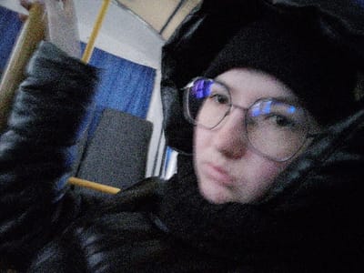 An individual is depicted in a close-up, low-light photograph taken inside what appears to be a public transport vehicle in Novyi Shagirt, Russia. The subject, who appears to be female, occupies the majority of the frame, looking directly towards the camera. She wears a dark, possibly black, beanie or knitted cap and large, light-rimmed eyeglasses with prominent blue reflections on the lenses. Her outerwear consists of a dark, puffy or quilted jacket/coat, with a dark scarf or high collar covering part of her lower face. Her expression is neutral.

The background elements are consistent with a passenger compartment. To the left and slightly behind the individual, a dark grey upholstered seat back is visible, featuring a horizontal wooden or light-colored bar, likely a handle or structural component. Further back, a textured blue surface, possibly upholstery or wall paneling, is discernible. A partial yellow vertical pole, common in public transport for standing passengers, is visible above the seat. The overall image exhibits significant digital noise, characteristic of a photograph taken in low light conditions.