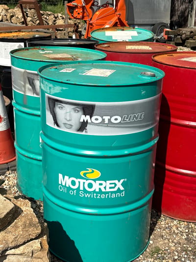 The image shows a collection of stacked metal barrels in an outdoor environment, likely in the Vale of White Horse, United Kingdom. The barrels are of varying colors; red, turquoise and black. The turquoise barrels are labeled with "MOTOREX Oil of Switzerland" and "MOTOLINE" branding. A tractor or similar machinery with orange parts is partially visible in the background, along with a pile of cut wood and rocks. The ground is covered in gravel and small stones.