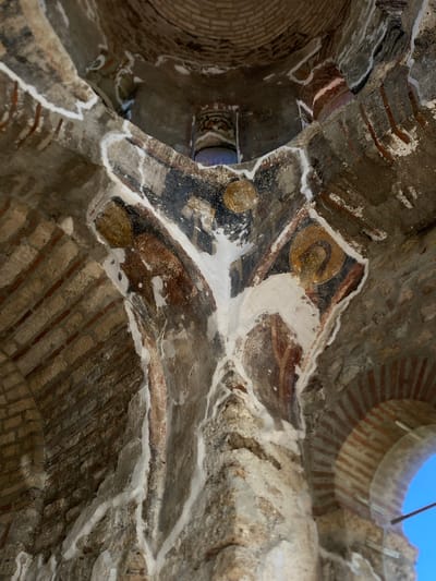 The image shows the interior of a domed structure, likely a church or monastery, with visible brickwork and faded frescoes. The frescoes depict figures with halos, suggestive of religious imagery. The structure appears old and possibly undergoing restoration, evidenced by the white plaster applied in some areas. The view is upward, capturing the apex and the curving walls. The environment is dimly lit, with the exception of light entering from an arched opening at the right, revealing a clear blue sky. The location is identified as Raska Municipality, Serbia.