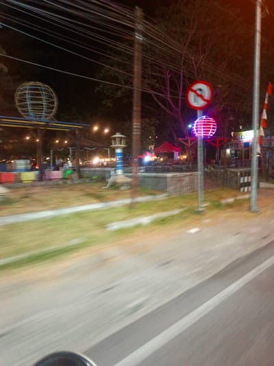 This is a motion-blurred, nighttime photograph taken from a moving vehicle in Luwoo, Indonesia. The foreground shows a grey asphalt road with white lane markings. To the left, a patch of blurred green grass is visible. In the mid-ground, there are several objects and structures. A large, wireframe globe illuminated with blue and purple lights stands prominently. To its left, another, larger wireframe globe structure, also illuminated, is visible. Behind these globes, there are various stalls and buildings, some with awnings and lights, suggesting an outdoor market or gathering place. Red and yellow rectangular structures are also present. A red tent is visible in the background. A traffic sign with a red circle and a diagonal line, prohibiting something, is positioned above the illuminated globe. A tall, slender pole with a red and white striped flag is to the right of the frame. The overall scene is dimly lit with artificial lights illuminating specific areas. The motion blur indicates a fast shutter speed or camera shake.