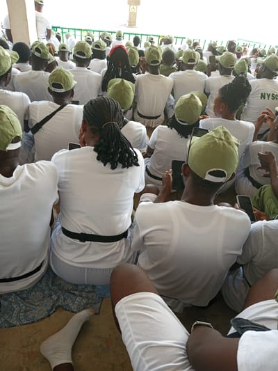 The image shows a group of young people, mostly men, wearing white t-shirts and light green caps, sitting on the ground. Some are looking at their phones. A young woman with braided hair is visible in the foreground. The scene appears to be outdoors, possibly at a National Youth Service Corps (NYSC) orientation camp in Ajiwa, Nigeria, given the common attire and the "NYSC" text on one of the shirts. The environment suggests a gathering or event where participants are waiting or relaxing. The lighting indicates it is daytime.