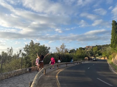 The image displays a curving asphalt road and an adjacent stone-paved sidewalk in Deià, Spain, observed under a blue sky with scattered white clouds. The road, featuring a solid white edge line and a broken white center line, extends into the distance curving towards the right. Five individuals are present on the left-hand sidewalk, walking away from the viewer. The closest person, from behind, has long dark hair, is wearing a white top and a brown patterned skirt, and has both arms raised and extended outwards. Behind this individual, a person in a light pink top and another in a bright pink top are visible. Further back on the sidewalk, a fourth person wears a white top and patterned skirt, and a fifth wears a blue patterned dress. The sidewalk is bordered by a low dry-stone wall and a wooden post-and-rail fence. The surrounding environment consists of lush green Mediterranean trees and bushes covering rolling hills. Several terracotta-roofed buildings are nestled among the foliage on the distant hills. A tall, dark, columnar tree stands prominently on the far right side of the road. In the distance on the right side of the road, a white SUV is parked or stopped, with two indistinct figures standing near it. The lighting suggests late afternoon or early evening.