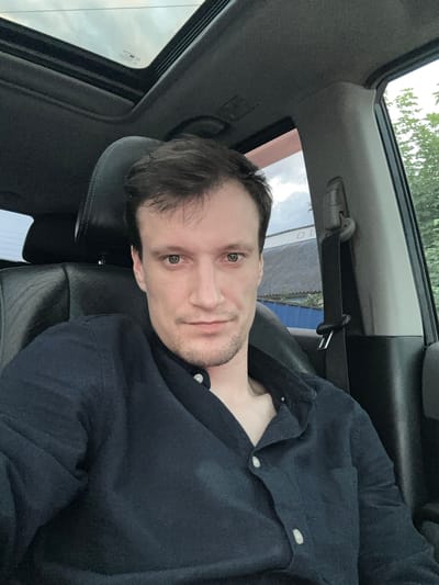 A man is sitting in the driver's seat of a car, looking at the camera. He is wearing a dark blue collared shirt. The car has a sunroof that is open, showing a cloudy sky. Outside the car, there are trees and a building with a corrugated roof. The image was taken in Shatava, Ukraine.
