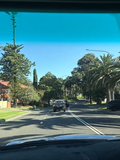 The image depicts a street scene viewed from inside a car. The road is lined with trees, casting shadows on the asphalt. A silver truck is driving ahead on the road. Buildings are partially visible on the side of the street. The sky is clear and blue, visible through the car's tinted windshield. There are double white lines on the road.