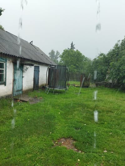 The image depicts a backyard scene in Sosnivka, Ukraine, during a heavy rain shower. A white building with a dark, corrugated roof and blue trim around a window is visible on the left. Two weathered, blue wooden doors are present on the side of the building. In the yard, a trampoline with a black safety net is set up. The ground is covered in green grass, with some patches of mud and scattered leaves. Behind the trampoline, a wooden fence and some overgrown vegetation can be seen. The sky is overcast and gray, indicative of the rainy weather. The rain is visible as streaks of water falling throughout the frame.