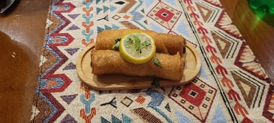 Two golden-brown, crispy rolls are arranged on a small, oval wooden serving board. A lemon slice sits atop the lower roll. Green sprigs of cilantro are tucked around the base and between the rolls. The board rests on a colorful table runner with geometric patterns in beige, brown, red, blue, and orange. The surrounding table surface is a warm brown.