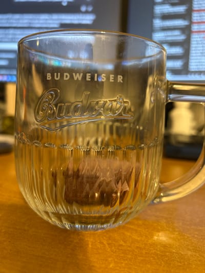 The image shows a close-up of a clear glass Budweiser mug resting on a wooden surface. Inside the mug, there is a small, brown object submerged in a clear liquid. The mug has vertical ridges around its base and a prominent Budweiser logo embossed on its side. The background is blurred, but appears to be a computer screen displaying text. The scene is lit by ambient light, creating reflections on the glass. The image was taken in the city of Slovenský Grob, Slovakia.