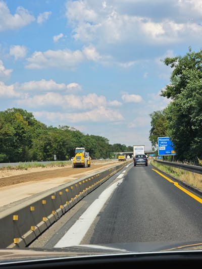 The image depicts a view from inside a vehicle traveling on a highway in Rüsselsheim am Main, Germany. On the left side of the highway, there is a construction zone with a yellow steamroller operating on a dirt surface. Concrete barriers with yellow reflectors delineate the construction area from the active lanes of traffic. On the right side of the highway, a blue road sign indicates "Rüsselsheim - Mitte, Trebur, Opel" and an exit at 100 meters. Another smaller blue sign displays "Ausfahrt 55". A dark blue car is traveling ahead in the right lane. The highway is bordered by trees and vegetation on both sides. The sky is blue with scattered white clouds.