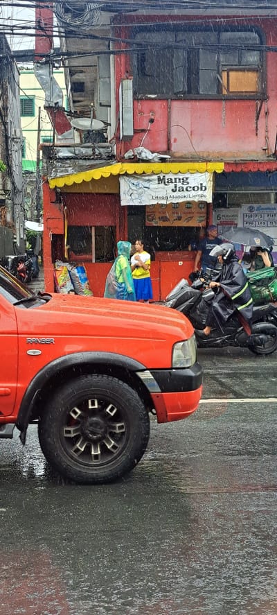 The image depicts a street scene in Mandaluyong City, Philippines, during a heavy downpour. In the foreground, the front wheel and fender of an orange Ford Ranger pickup truck are visible. The truck is parked on a wet asphalt road, with raindrops clearly visible in the air and on the vehicle's surface.

Behind the truck, a red building with a yellow awning serves as a storefront. A sign above the awning reads "Mang Jacob" with text indicating "Litson Manok/Liempo" (roast chicken/liempo). Below this, a price list is partially visible.

Several people are present in front of the storefront. Two individuals are wearing raincoats; one in bright yellow and the other in blue. A young girl in a yellow shirt and blue skirt stands between them, appearing to converse.

Further to the right, a person wearing a black raincoat rides a black motorcycle. This person is holding a black umbrella. Another individual, partially obscured by the motorcycle and umbrella, is also visible. Bags of what appear to be groceries are stacked near the storefront.

The environment is characterized by the continuous rain, wet surfaces, and a chaotic arrangement of electrical wires overhead, typical of urban Philippine settings. The overall impression is one of everyday life continuing despite adverse weather conditions.