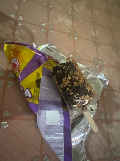 A half-eaten chocolate-covered ice cream bar with nuts on a stick lies on its foil wrapper. The wrapper, which is partially open, reveals yellow and purple branding with Cyrillic text. The ice cream is resting on a light pink tiled surface with a faint heart-shaped pattern. The scene appears to be indoors, on a floor or table. The lighting is dim. There is no indication of the location being in the city of Aqköl, Kazakhstan.