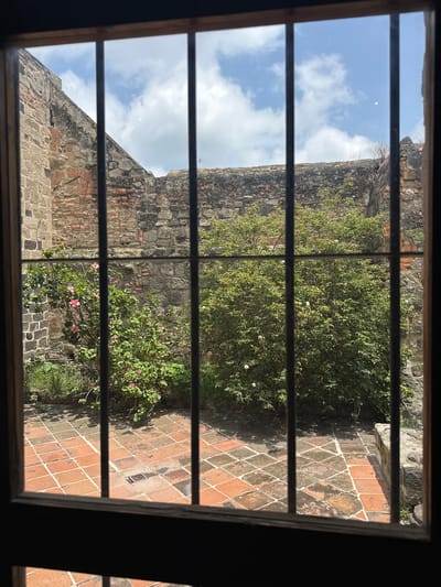 The image shows a courtyard viewed through a window with dark wooden frames and metal bars. The courtyard is paved with terracotta tiles, some of which are worn or stained, and there is a drain grate in the center of the paved area. On the left, there is a wall constructed from stone and brick, with some areas of exposed brick and mortar. Adjacent to this wall, a flowering bush with pink blossoms is visible. To the right of this bush is a denser, green shrub with small white flowers. The background consists of more brick and stone walls, suggesting an enclosed or partially enclosed space. Above the walls, a partly cloudy blue sky is visible. The scene appears to be an outdoor space within a historical building. The location is the former Augustinian convent in Huejotzingo, Mexico. There are no people visible in the image.