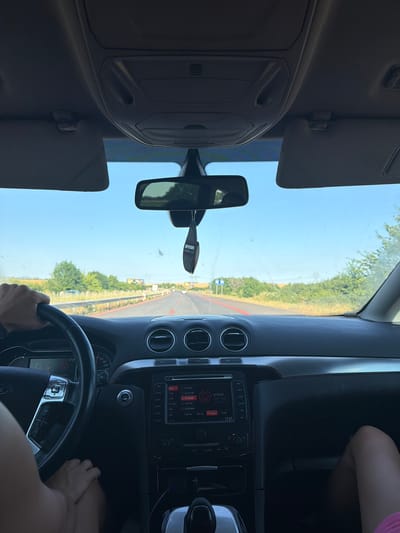 A first-person perspective shot shows the interior of a car driving on a highway. The road is paved and appears to be in good condition, with a red line marking the right edge. Trees and fields line both sides of the road, suggesting a rural or suburban environment. The sky is clear and blue. In the foreground, a person's hands are visible on the steering wheel, indicating they are driving. The car's dashboard and center console are visible, with a radio display showing station information and a clock. A black air freshener hangs from the rearview mirror. The image is taken during daylight. The location is identified as the highway near the city of Vinarsko, Bulgaria.