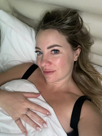 A woman with blonde hair and blue eyes lies in bed. She's facing the camera with a slight smile. She is wearing a black strap, possibly from a bra or tank top. Her hand rests on a white pillow or blanket, adorned with several rings and her nails are painted with a light color and glitter. The background includes a beige curtain or wall.