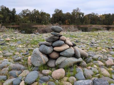 A stack of rounded rocks forms a small cairn on a riverbank. Various shades of grey and brown stones are piled together, surrounded by other scattered rocks and sparse patches of green grass. The background features a calm river, reflecting the sky, with a line of trees along the opposite bank. The sky is overcast, casting a soft light on the scene.