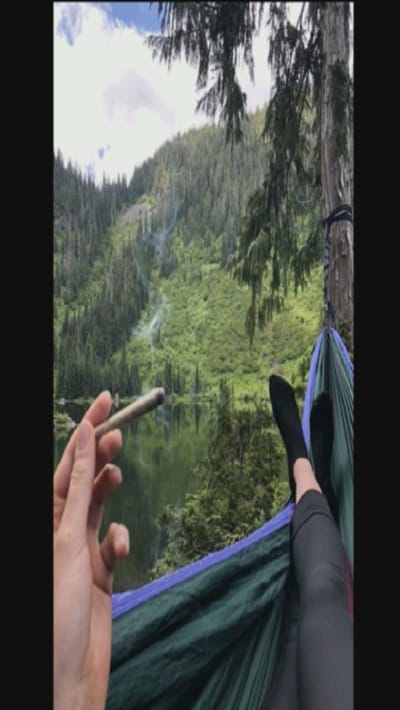 A person lies in a hammock with their feet up, wearing black socks and leggings. Their hand holds a rolled cigarette. In the background is a lake surrounded by lush green trees. The sky is cloudy, and there are hints of smoke on the mountainside in the distance.