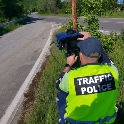 A traffic police officer, identified by the "TRAFFIC POLICE" inscription on their bright yellow vest, is operating a blue radar speed detection device positioned along a road. The location is indicated by a sign in the background, showing this is Balkantsi, Bulgaria. The officer is wearing a navy blue baseball cap, long-sleeved shirt, and pants. A black smartwatch is visible on their left wrist. The speed detection device is connected by cables, and the officer appears to be looking at the screen. The road is adjacent to a grassy area and some trees. A rusty metal guardrail is visible along the roadside.