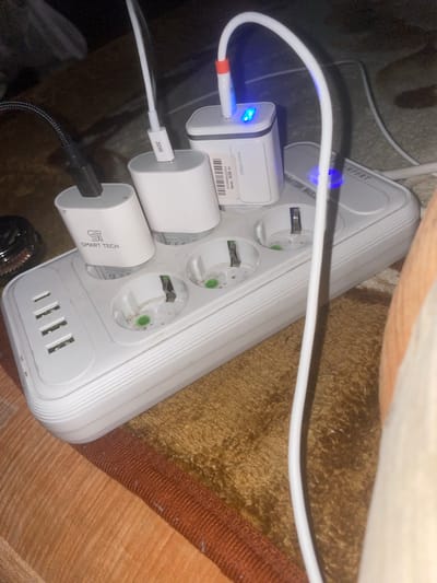 The image displays a white power strip on a brown and orange textured surface, likely a carpet. Several white and black power adapters are plugged into the power strip. One white adapter has "30W" printed on it and is connected to a white cable. Another white adapter is labeled "SMART TECH" and is connected to a black cable. A third white adapter, larger than the others, has a blue LED light indicating it is active and is connected to a white cable with a red connector. The power strip also has multiple USB ports, four of which are visible. The environment appears to be indoors and dimly lit. The location of Панфилово, Kazakhstan is not depicted or suggested by any visual elements in the image.
