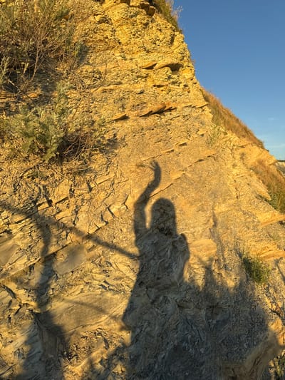 The image depicts a sunlit cliff face with stratified rock layers and sparse vegetation. A shadow of a person with one arm raised is cast prominently on the rock. The cliff is a mix of yellow and grey rock, with dry grass and small green plants growing from crevices. The sky above is a clear blue. This scene is located in the vicinity of Varvarovka, Russia.