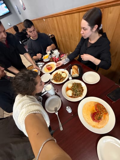 The image captures a dining scene at a restaurant in Des Plaines, Illinois. A group of people are seated around a dark wood-grained table laden with food. A woman is serving food to another person at the table. Dishes include stuffed tomatoes and a mushroom-based dish. Other items such as bread, beverages, and condiments are visible. The restaurant has wood paneled walls, and the atmosphere is casual.