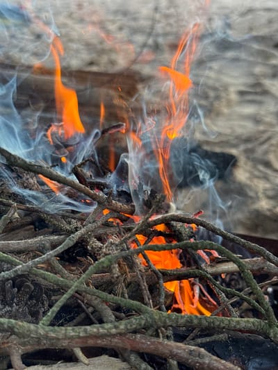 The image shows a close-up view of a small campfire. Dry twigs and branches are piled together and burning, producing orange flames and wisps of white and grey smoke. In the foreground, several thick, moss-covered branches are visible, obscuring part of the fire. The background is blurred and appears to be a rough, light-colored surface, possibly wood or stone. The scene is set in an outdoor environment. The location of Fort Krasnaya Gorka in Russia is not visually represented in this image.