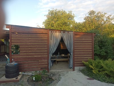 The image shows an outdoor covered seating area or gazebo. The structure is made of dark brown wooden planks with lighter horizontal slats. A grey fabric curtain hangs across the entrance, which is open. Inside, a wooden table and benches are visible, with some items on the table, including a bottle and what appear to be plates with food. The ground inside the gazebo is covered in gravel.
To the left of the gazebo, there is a unique outdoor sink constructed from stacked car tires. A faucet is attached to the top tire, and a blue brush rests on the edge. A garden hose is coiled nearby. A framed picture of yellow roses hangs on the wooden wall to the left.
The scene is set in a garden environment, with lush green trees and bushes visible behind and around the structure. Ferns are prominent in the foreground on the right. The ground outside the gazebo is a mixture of concrete and gravel paths. The lighting suggests it is late afternoon or early evening, with the sun casting a warm glow on the trees. The sky is partly cloudy. There are no people visible in the image. The overall impression is of a rustic, functional outdoor living space.